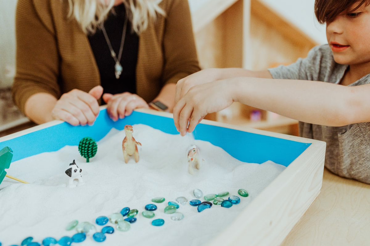 A child places a figure in a sandtray while a therapist sits beside him in a Beyond Healing play therapy room