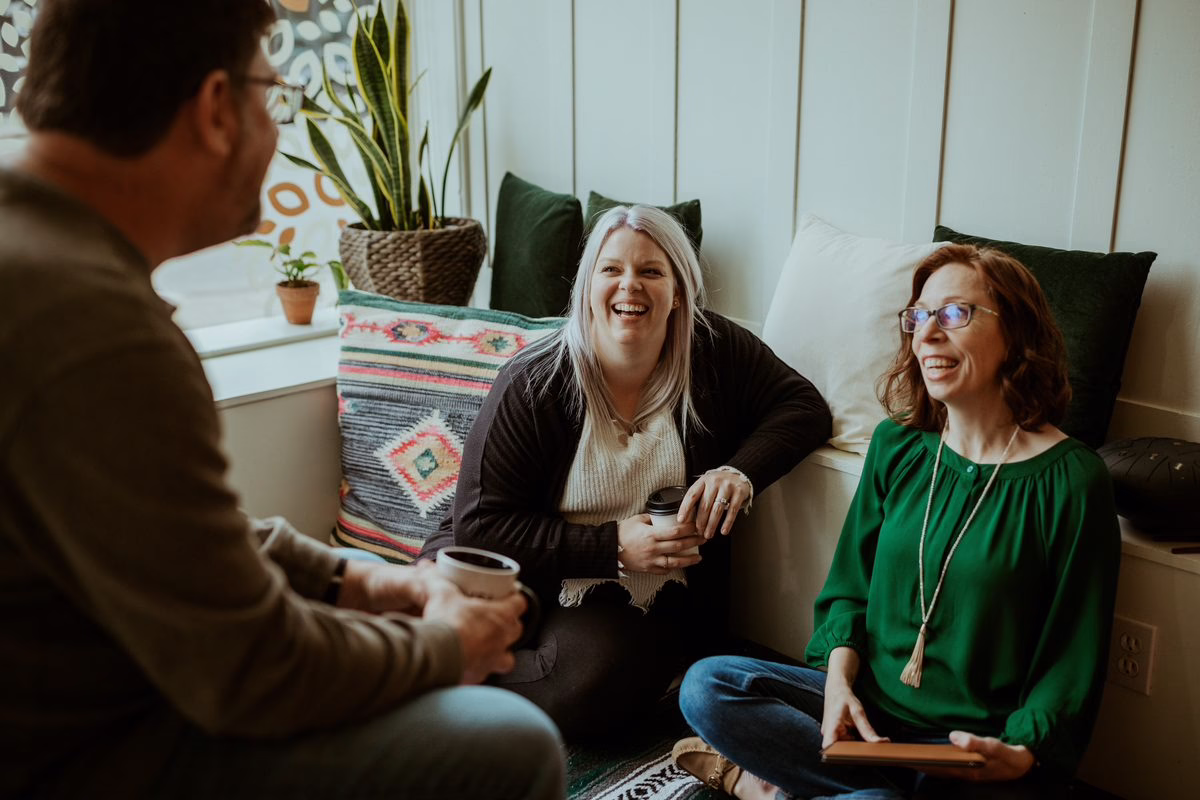 Three people laughing together over coffee in a sunlit Beyond Healing office